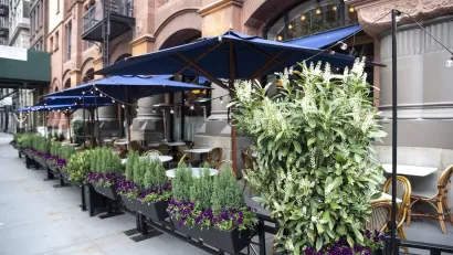 Outdoor dining patio with tables and boxes of flowers surrounding it on New York City sidewalk.