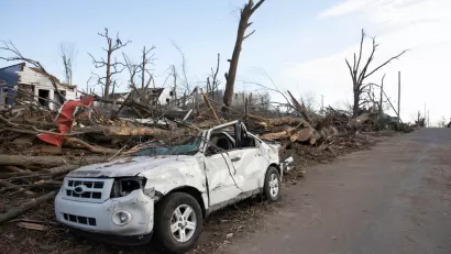 Heavily damaged white small SUV parked on street with heavy tornado damage and leveled houses in Mayfield, Kentucky.