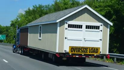 Tan modular building with white swinging barn/garage door being transported on freeway with yellow "Oversize Load" banner across back.