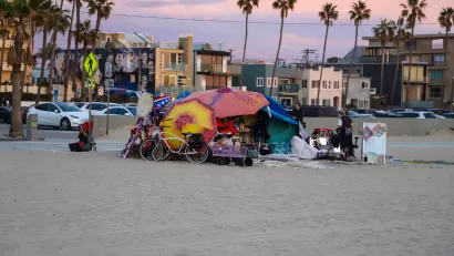 Colorful tents built to shelter unhoused people on beach at Venice Beach, California with apartment buildings and palm trees in background against a purple sunset sky