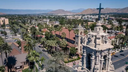 Aerial view of Riverside, California with ornate Catholic church tower in foreground and mountains in the distance.
