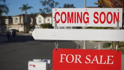 Close-up on red and white "Coming Soon" and "For Sale" signs in front of single-family homes on sunny day.