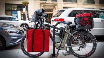 Black e-bike with red food delivery bags locked at bike rack on New York City street.