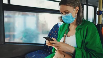 Woman in green sweater wearing light blue medical mask sitting on bus looking at her phone.