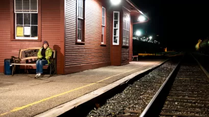 Womanin yellow jacket sitting on bench at outdoor train station with red wood depot building at night.