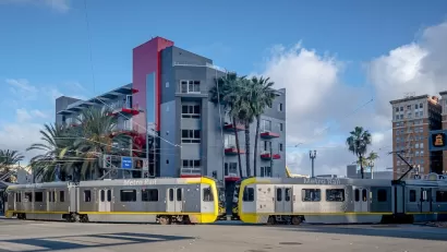 Two silver trains leaving station in Long Beach, California in front of mid-rise residential building.