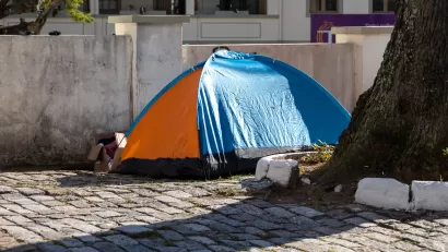 A orange-and-blue tent set up next to a tree in a paved courtyard and a cardboard box of posessions next to the tent entrance.