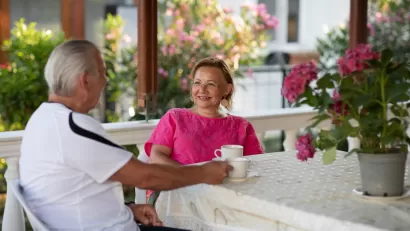 Elderly woman in pink shirt sitting at table on porch across from elderly man in white shirt. A pot of pink flowers sits on the table.
