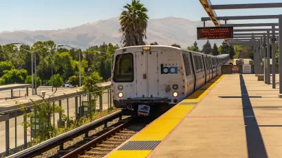 San Francisco Bay Area Rapid Transit train at elevated station with hills and palm tree in background.
