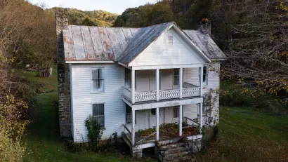 White two-story abandoned farmhouse in lush green field surrounded by trees in West Virginia.