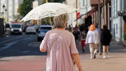 Older woman walking on city street holding white lace sun parasol.