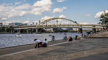 People on bikes and sitting on ledge on Pittsburgh riverfront with arched bridge in background