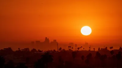 Sun rising over downtown Los Angeles with tall palm trees visible in foreground. Image is bright orange-red indicating extreme heat.