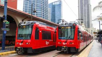 Two red San Diego trolley cars at Santa Fe Depot station with high-rise buildings in background.