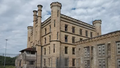 Deralict stone prison building with turrets and a chain link fence in foreground, under a gray, cloudy sky.