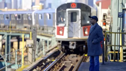 Subway train pulling into elevated outdoor platform in New York City with one passenger waiting.