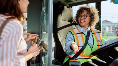 Female bus driver wearing safety vest sits in driver's seat, talking with woman passenger boarding bus