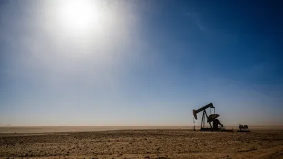 Distant view of oil derrick on barren plain with harsh sun in background.