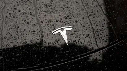 Close-up on silver Tesla logo on black car hood dotted with raindrops.