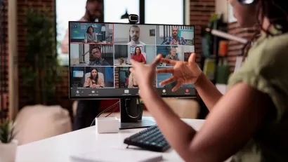 Person wearing headphones sitting at desk with monitor showing other people on Zoom meeting.