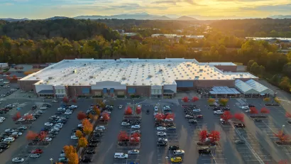 Aerial view of large parking lot in front of large grocery or big-box store with small fall-colored trees among parked cars and sunset sky in background.