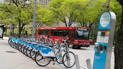 Blue and white Pittsburgh bike share bikes lined up at a station with a red city bus on street in background.