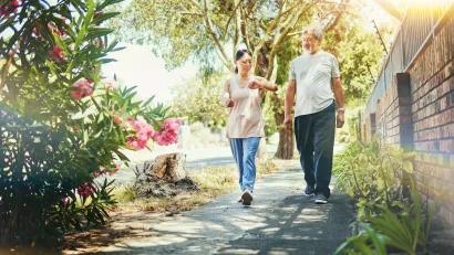 Older man and woman walking down sidewalk landscaped with bougainvillas next to a brick wall on a sunny day.