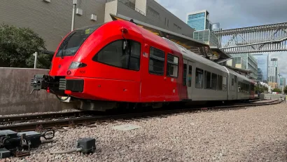 Red modern train on tracks with large warehouse-type building behind it in Austin, Texas.