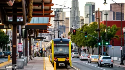 Yellow and black Metro light rail train pulling up to outdoor platform with Los Angeles skyline in background.