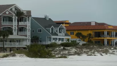 Three colorful, large beachfront homes, one khaki, one blue, and one yellow, with a small dune in front and flat sand in foreground.