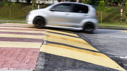 Small silver car driving over wide soeed cushion on asphalt road.