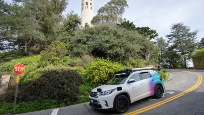 White self-driving car at stop sign coming down hill with Coit Tower in background in San Francisco, California.