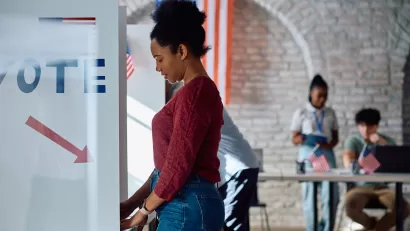 A side view of a young Black woman in a red sweater and jeans standing at a ballot box in a gymnasium with white walls and an American flag in the background.