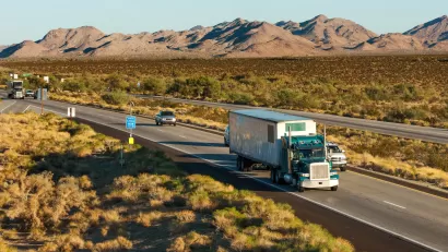 Double-lane interestate in the desert with two semi-trucks and five cars driving toward the camera, low mountains in the background.