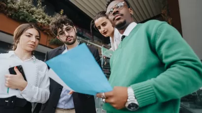Group of four young professional people standing in front of urban building looking at documents.
