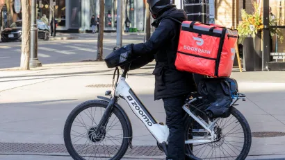 Person in black clothing and helmet with red DoorDash box on back of electric bike on urban street.