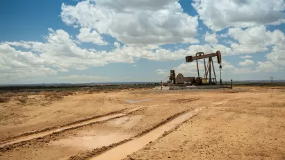 Distant view of brown oil pump jack in a flat desert with cloudy blue sky.