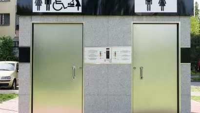 Outdoor public restrooms with two silver doors and icons for men, women, wheelchair accessibility, and baby changing above doors.