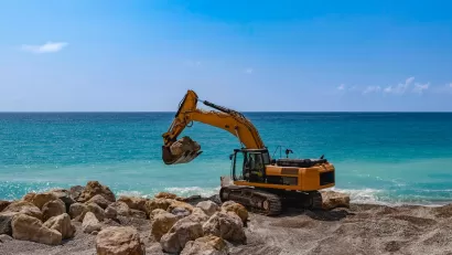 Yellow excavator on rocky beach with bright blue water in background.