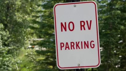 Close-up of white sign with red text 'No RV parking' against green foliage.