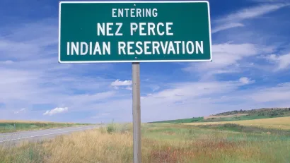 Green roadside sign with white text reading "Entering Nez Perce Indian Reservation" against grassy field and blue sky.