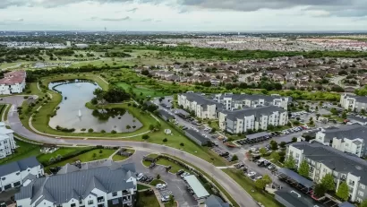Aerial view of suburban mixed-use development outside Austin, Texas with lake in foreground and complex of apartment buildings.