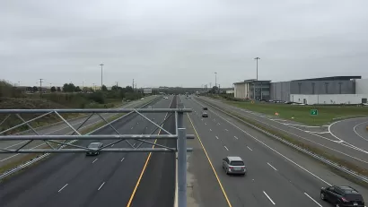 A wide highway with a few cars on a gloomy day. Low-rise commercial buildings and more car infrastructure is visible to the side of the road.