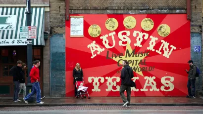 A woman with a baby in a stroller and two men are milling around in front a sign that reads Austin Texas: Live Music Capital of the World.