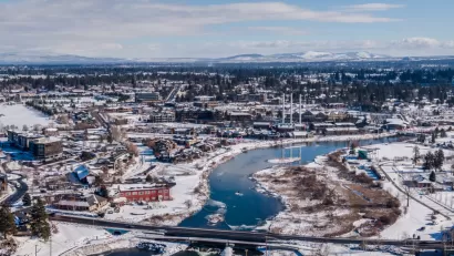 An aerial image of Bend, Oregon, covered in snow in the winter. The Deschutes River cuts through the middle of town, below an old mill.