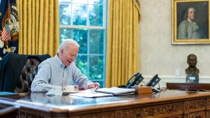 President Jose Biden appears in a collared, button down shirt while signing papers while seated at the desk in the Oval Office of the White House