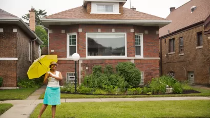 A Black woman stands in front of a home holding an umbrella and smiling.