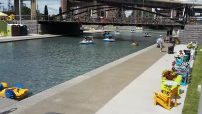A canal under a bridge in Buffalo is lined by chairs and tables, people walking about, looking at peddle boats on the water.