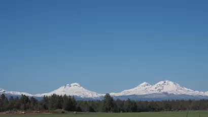 The Three Sisters, snow-covered mountains in Central Oregon, are visible in the distance beyond a farm.