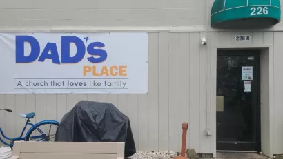 White sign on side of tan building with metal siding and green overhang, reading "Dad's Place, a church that loves like family."